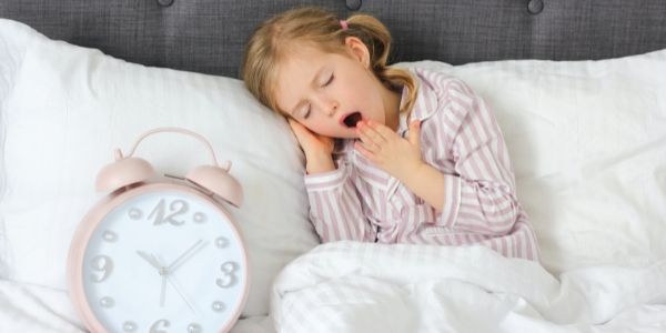 little girl yawning in bed with a big pink clock side her