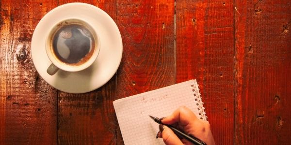 person writing in notebook on a red wooden table with a cup of coffee to the side